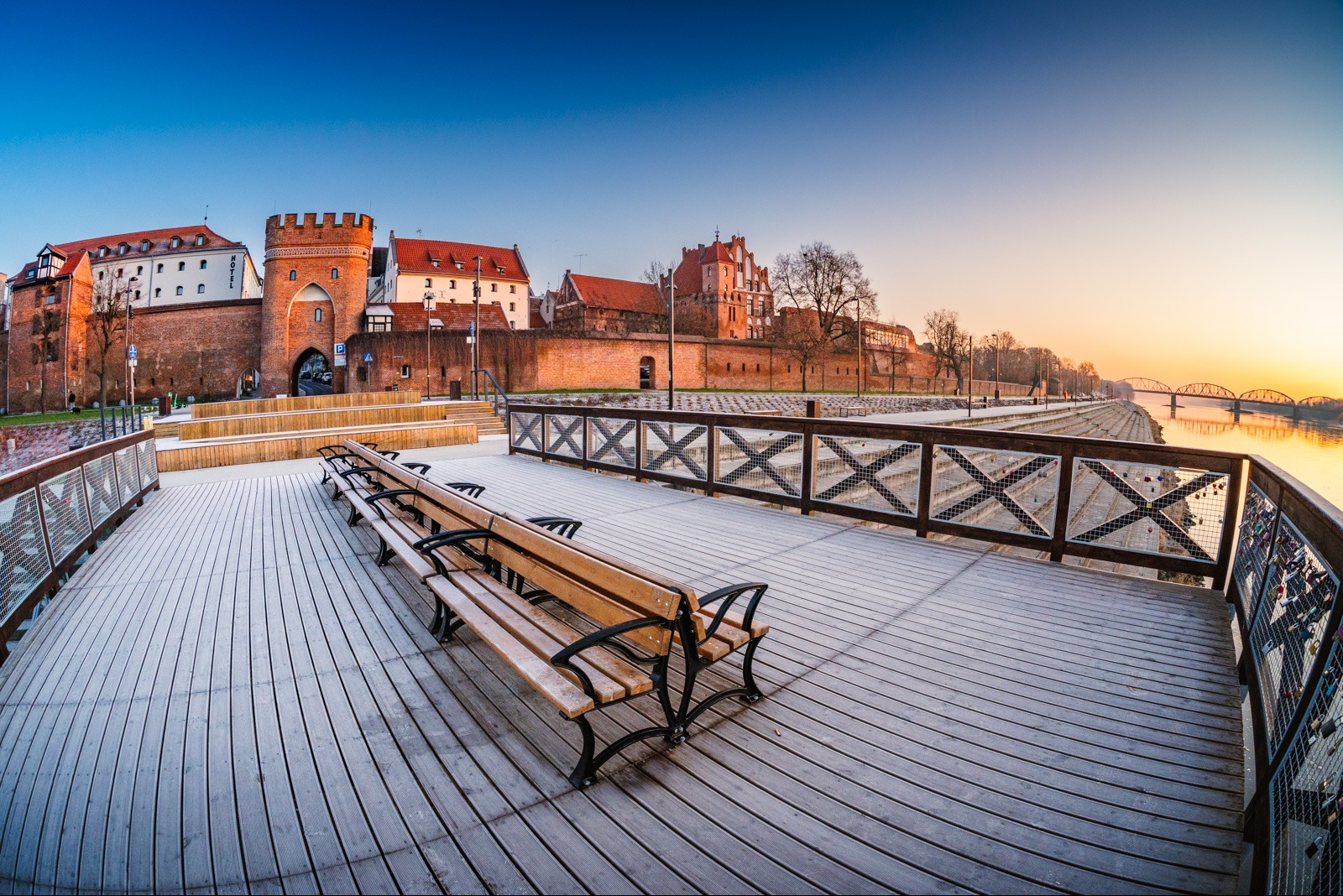 Panorama Torunia o świcie z perspektywy drewnianego molo z ławkami. W tle brama miejska, mury obronne i most na Wiśle w promieniach wschodzącego słońca.