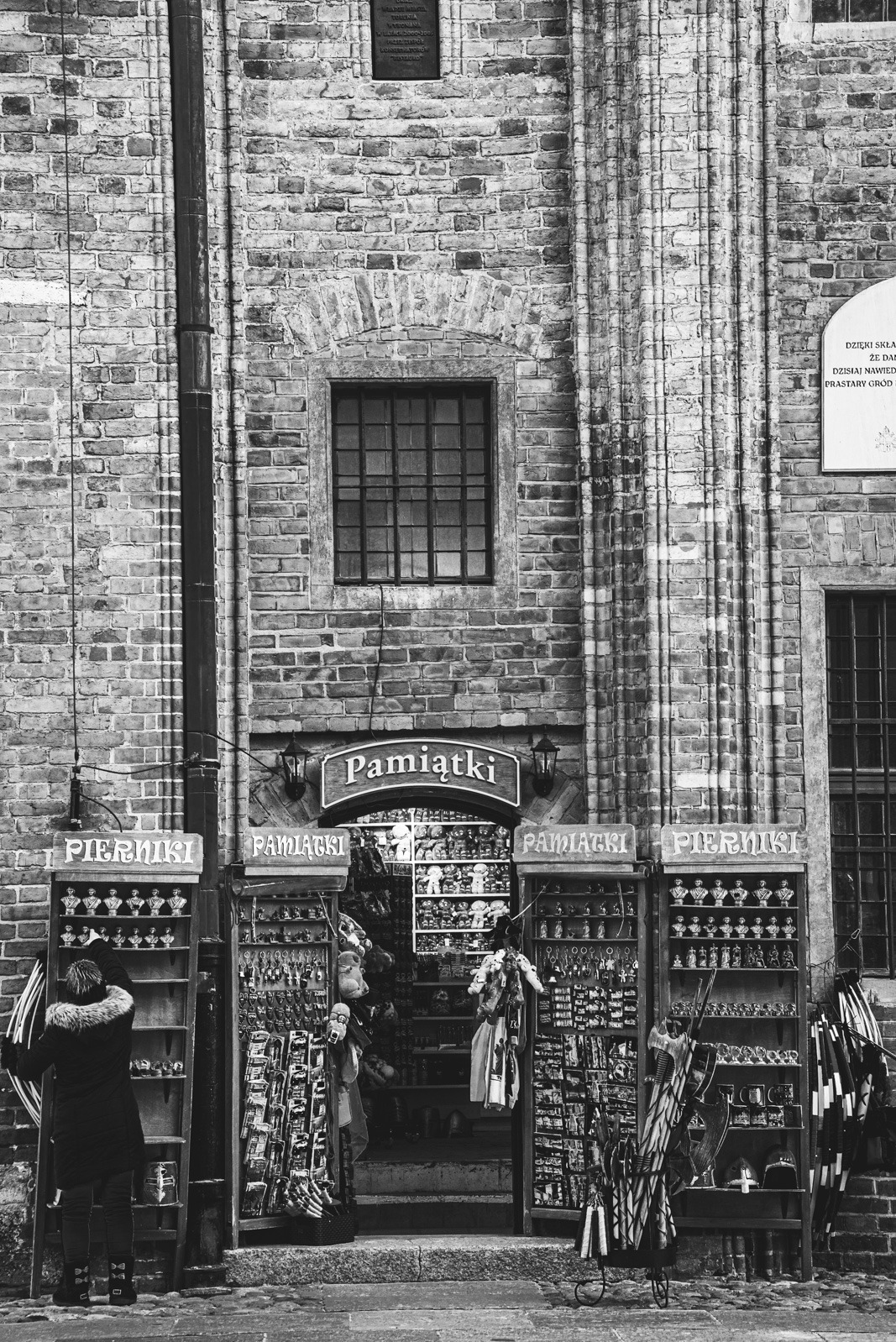 Czarno-białe zdjęcie fasady sklepu z pamiątkami w zabytkowej kamienicy. Widoczne regały z pamiątkami, okno i fragment tablicy informacyjnej. Osoba przygląda się towarom.