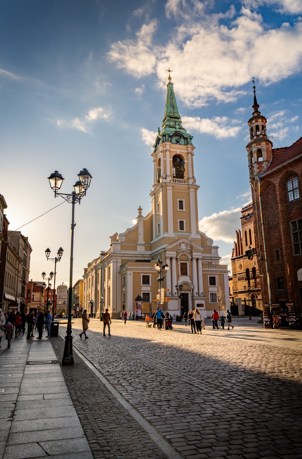 Słoneczny kadr z perspektywy ulicy na kościół z zieloną wieżą w Toruniu. Widoczni przechodnie i latarnie. Ciepłe światło podkreśla architekturę.