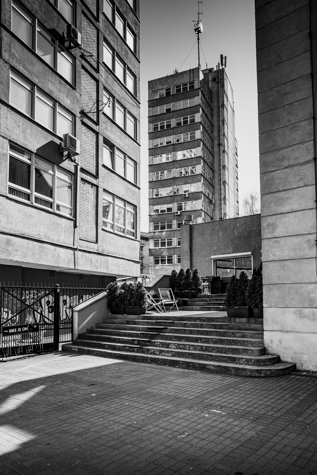 Czarno-białe zdjęcie miejskiej przestrzeni z budynkami, schodami, roślinami w donicach i metalową bramą z graffiti. W tle wysoki budynek z anteną.