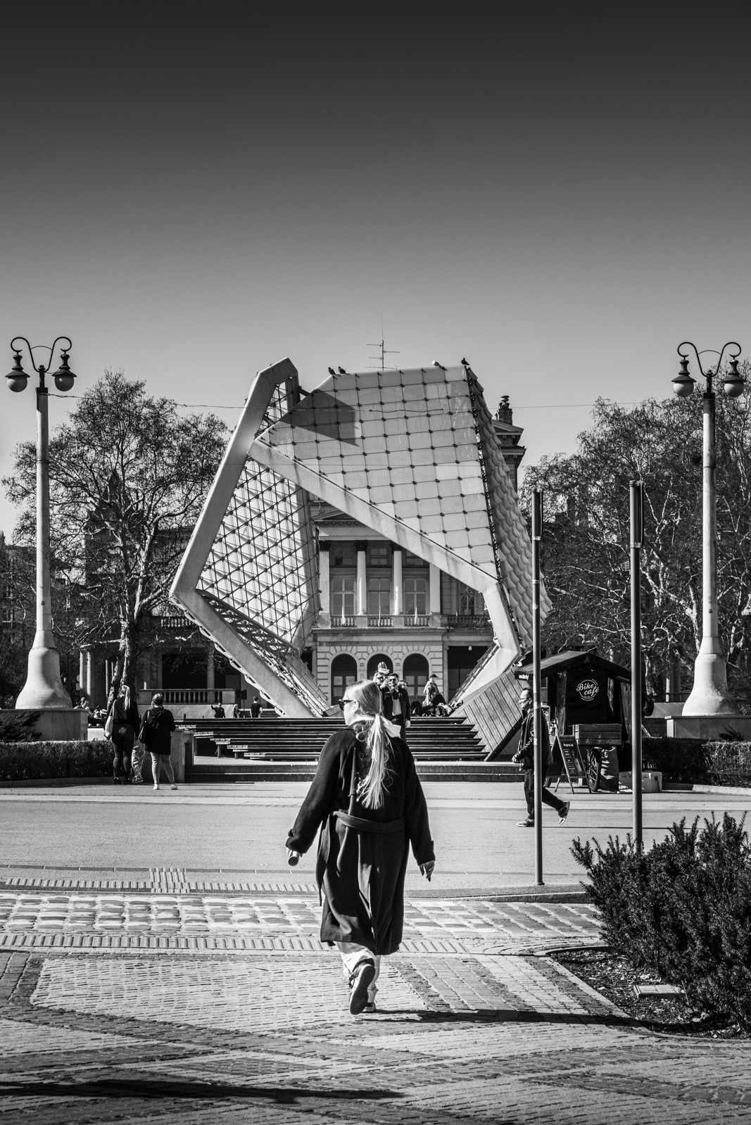 Czarno-białe zdjęcie kobiety idącej w stronę nowoczesnej fontanny na tle klasycystycznego budynku. Ujęcie z perspektywy pieszego, w słoneczny dzień.
