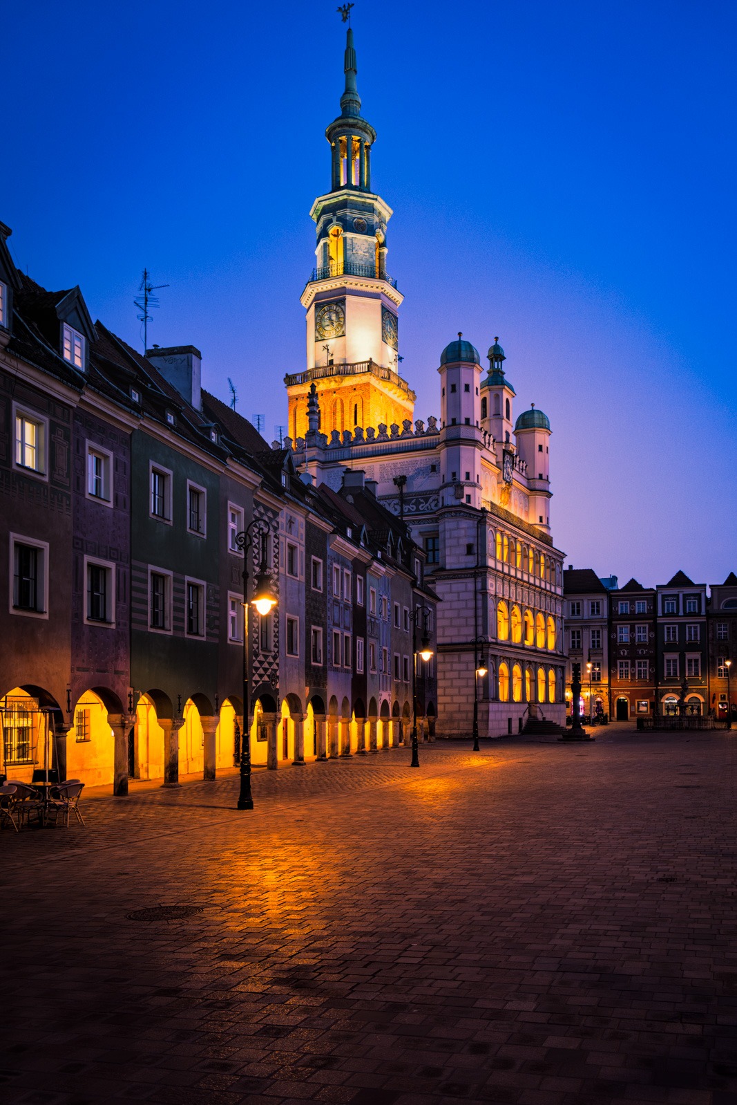 Nocny widok na Stary Rynek w Poznaniu. Oświetlony renesansowy Ratusz z wieżą zegarową góruje nad kolorowymi kamienicami z podcieniami. Puste, wybrukowane plac.
