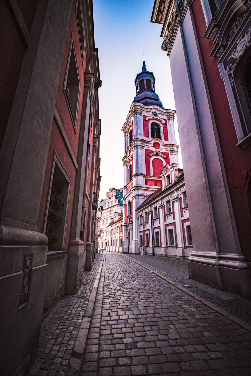 Ujęcie z dołu na barokowy kościół z czerwoną fasadą i wieżą, widziany z brukowanej uliczki w Poznaniu, Polska. Architektura miejska w ciepłych barwach.