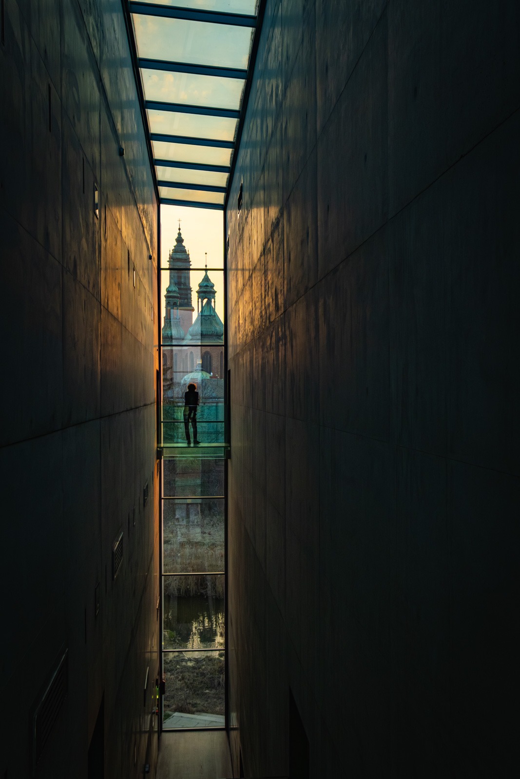 Wąski korytarz z przeszklonym dachem i podłogą, przez które widać sylwetkę osoby i fragmenty architektury miejskiej w ciepłym świetle zachodzącego słońca.
