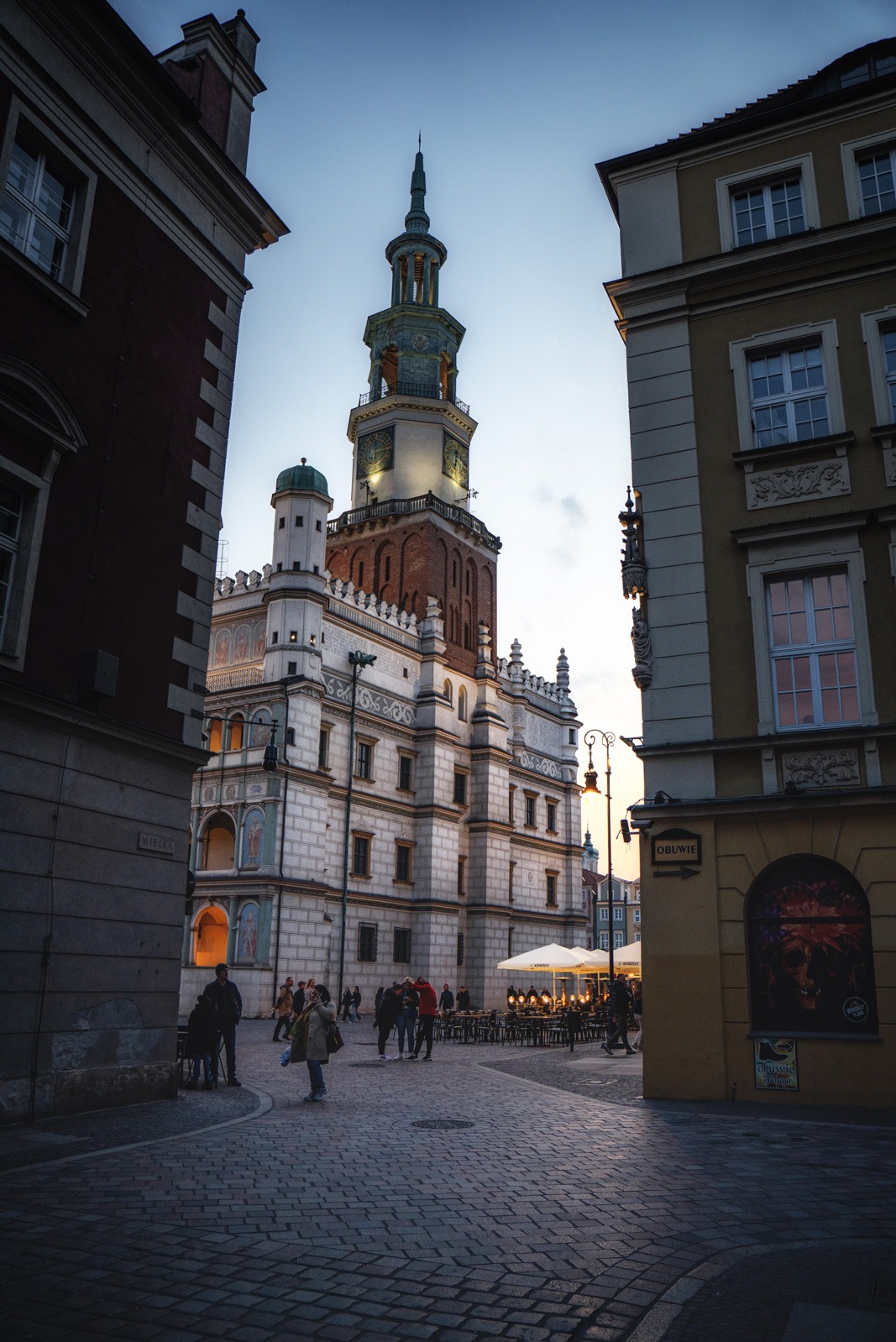 Rynek w Poznaniu o zmierzchu. Widok na renesansowy Ratusz z wieżą, otoczony kamienicami i spacerującymi ludźmi. Uliczne oświetlenie i parasole kawiarniane.