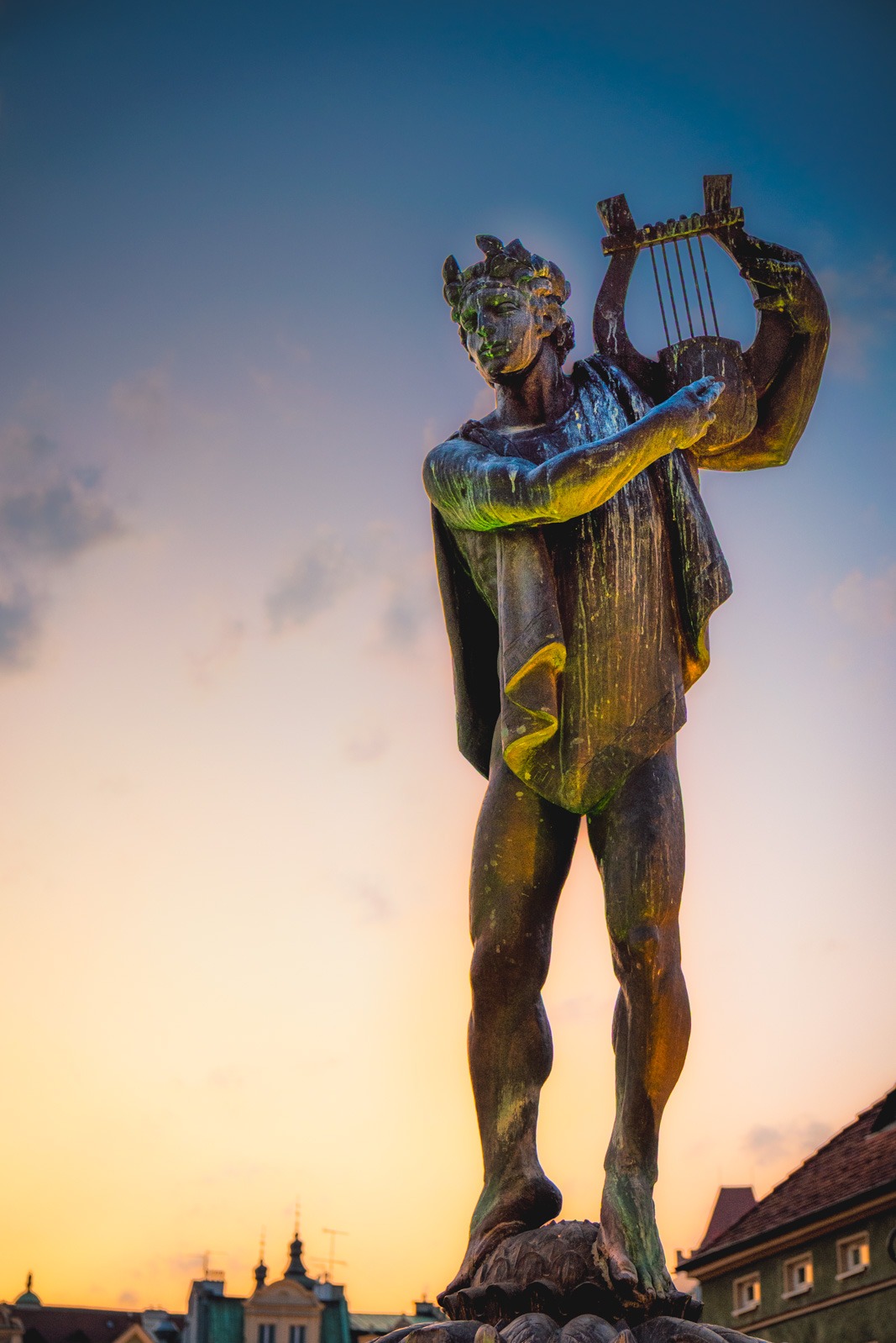 Statua mężczyzny z lirą na tle nieba o zachodzie słońca. Niskie ujęcie podkreśla monumentalność rzeźby i ciepłe barwy zachodzącego słońca.