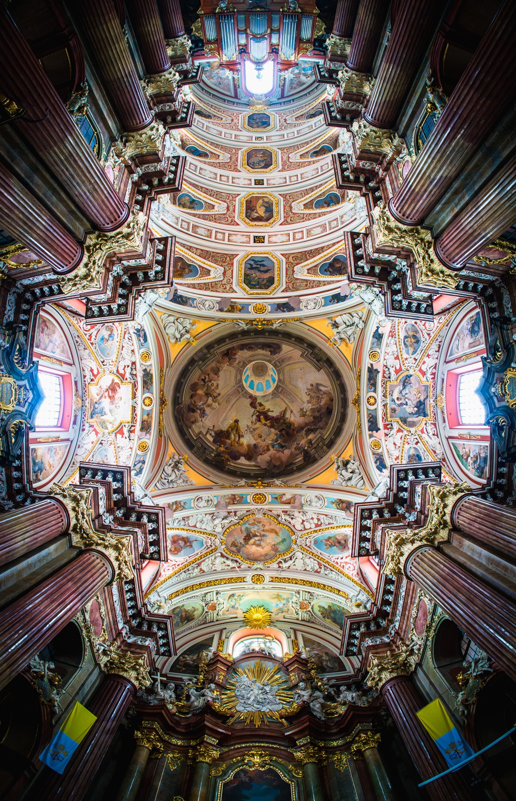 Barokowe wnętrze kościoła z bogato zdobionym sufitem, kolumnami i freskami, widziane z perspektywy żabiej. Intensywne kolory i detale architektoniczne.