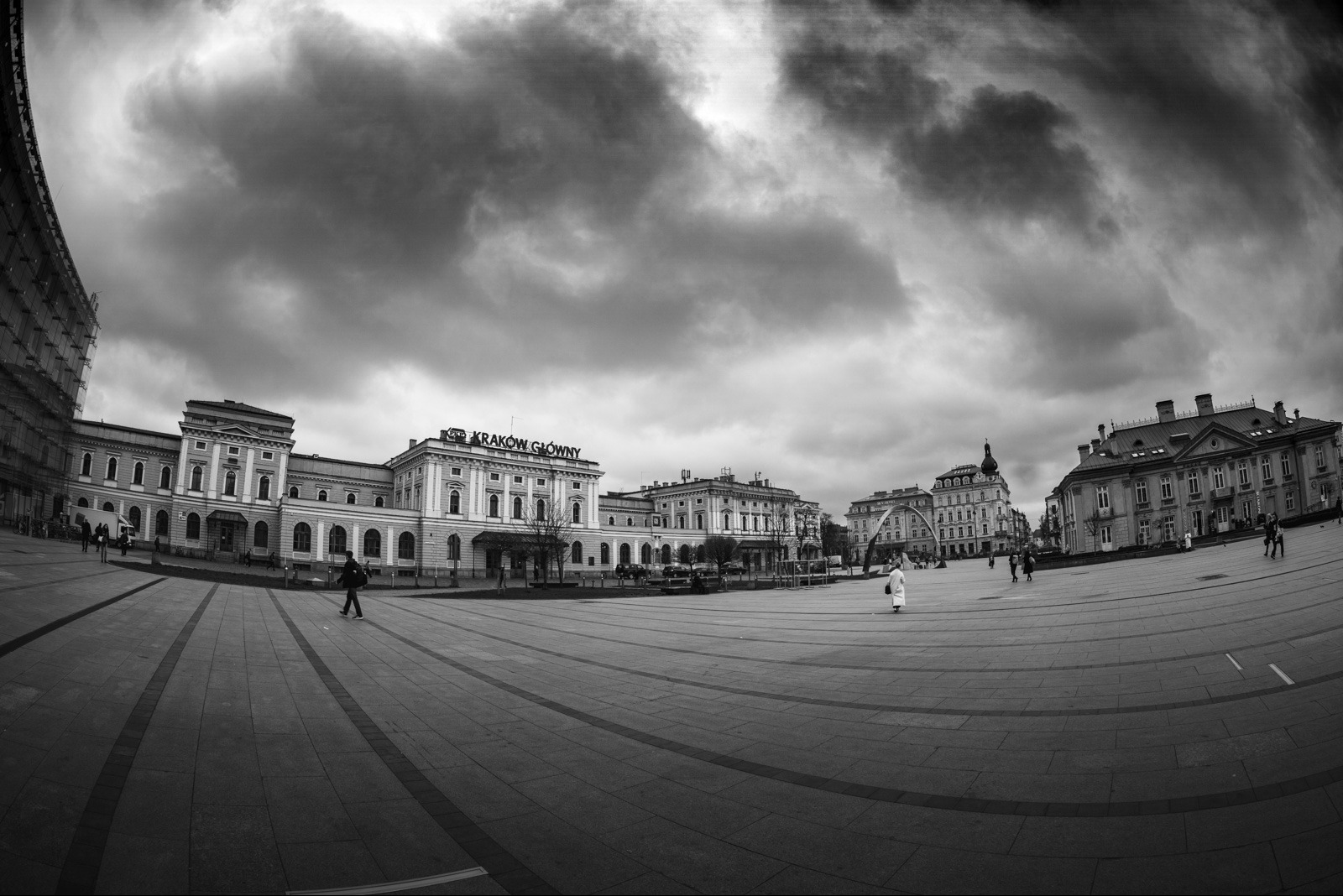 Czarno-białe zdjęcie z szerokim kątem: budynek Dworca Głównego w Krakowie pod pochmurnym niebem. Widoczny plac z przechodzącymi ludźmi i charakterystyczna architektura.