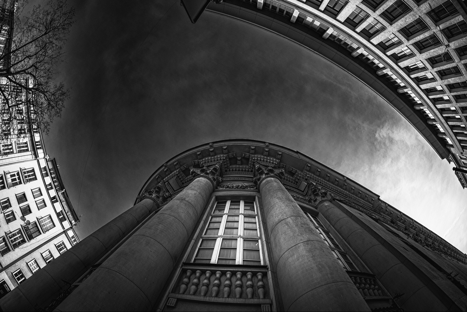 Monochromatyczne ujęcie z dołu na budynek z kolumnami, z widocznym fragmentem nieba i innymi budynkami. Perspektywa szerokokątna podkreśla architekturę.