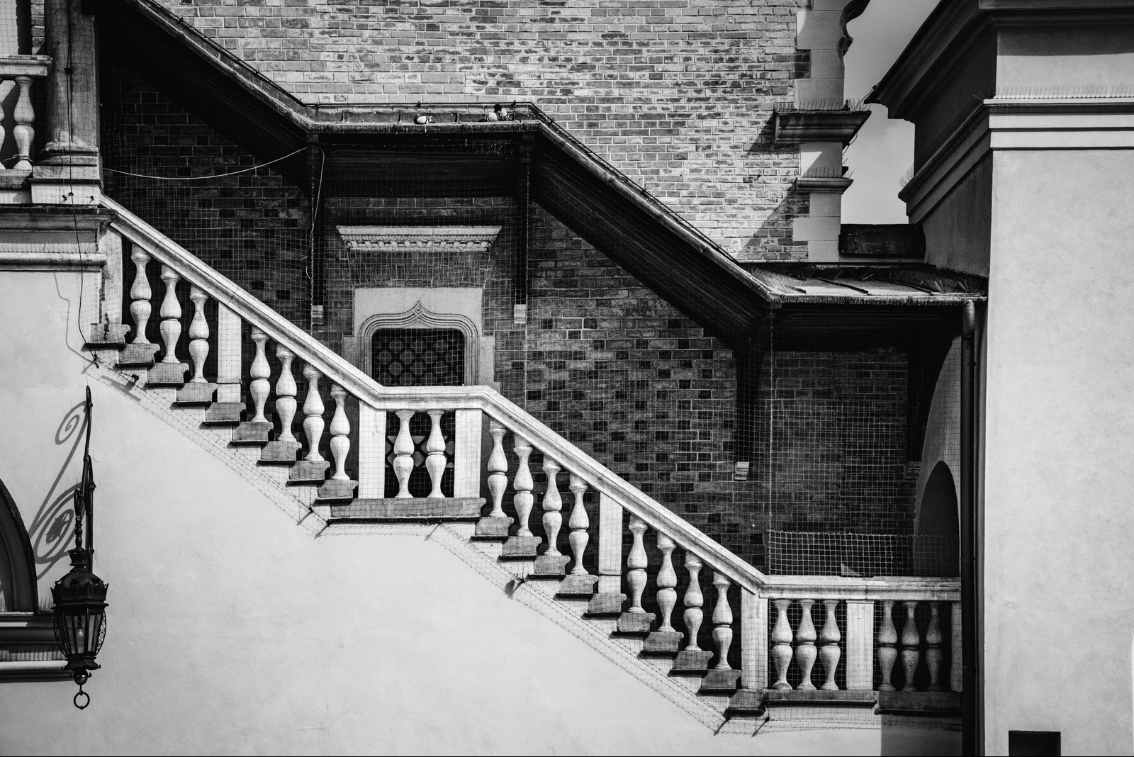 Czarno-białe zdjęcie zewnętrznych schodów z balustradą, prowadzących wzdłuż ceglanej ściany z oknem. W tle widoczny fragment budynku z elementami architektonicznymi.