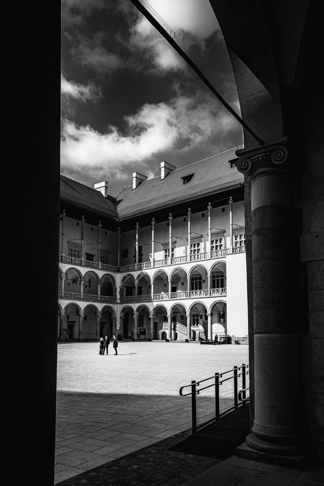 Czarno-białe zdjęcie dziedzińca zamkowego z arkadami, kolumnami i trzema osobami w oddali, uchwycone zza filaru, z widocznym fragmentem nieba z chmurami.