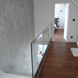 Gold Design Usługi remontowe - Korytarz z drewnianą podłogą i szklaną balustradą, ściana pokryta tapetą w odcieniach szarości, widok na otwarte drzwi do sypialni.