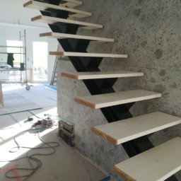 Gold Design Usługi remontowe - Nowoczesne schody z białymi stopniami i czarną, zygzakowatą konstrukcją nośną, w trakcie montażu przy ścianie wykończonej szarym kamieniem. W tle widoczne narzędzia i elementy wyposażenia wnętrza.