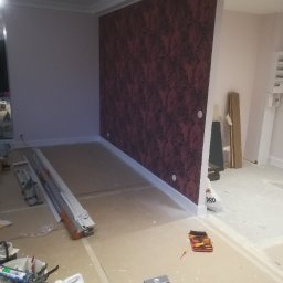 Gold Design Usługi remontowe - Pokój w trakcie prac wykończeniowych: ściana pokryta bordową tapetą z wzorem, listwy przypodłogowe, rozłożone narzędzia i materiały budowlane na podłodze, widoczne kable wystające ze ściany.