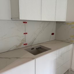 Gold Design Usługi remontowe - Biała kuchnia w trakcie montażu: marmurowe płytki na ścianie z czerwonymi krzyżykami dystansowymi, blat z wbudowanym zlewem i białe szafki wiszące.