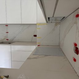 Gold Design Usługi remontowe - Białe płytki imitujące marmur na ścianie w kuchni, z czerwonymi klipsami poziomującymi. Widoczna płyta indukcyjna i białe szafki.