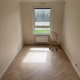 Gold Design Usługi remontowe - Jasne panele ułożone w jodełkę, kremowe ściany z listwami przysufitowymi, okno z widokiem na zieleń, złoty żyrandol. Krzesło zabezpieczone folią. Nowoczesne wykończenie wnętrza.