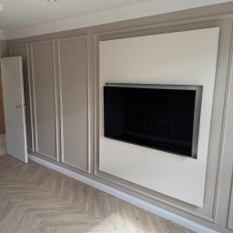 Gold Design Usługi remontowe - Eleganckie wykończenie salonu: ściana z dekoracyjnymi panelami w odcieniach beżu, wbudowany telewizor, parkiet w jodełkę, otwarte drzwi. Klasyczny design.