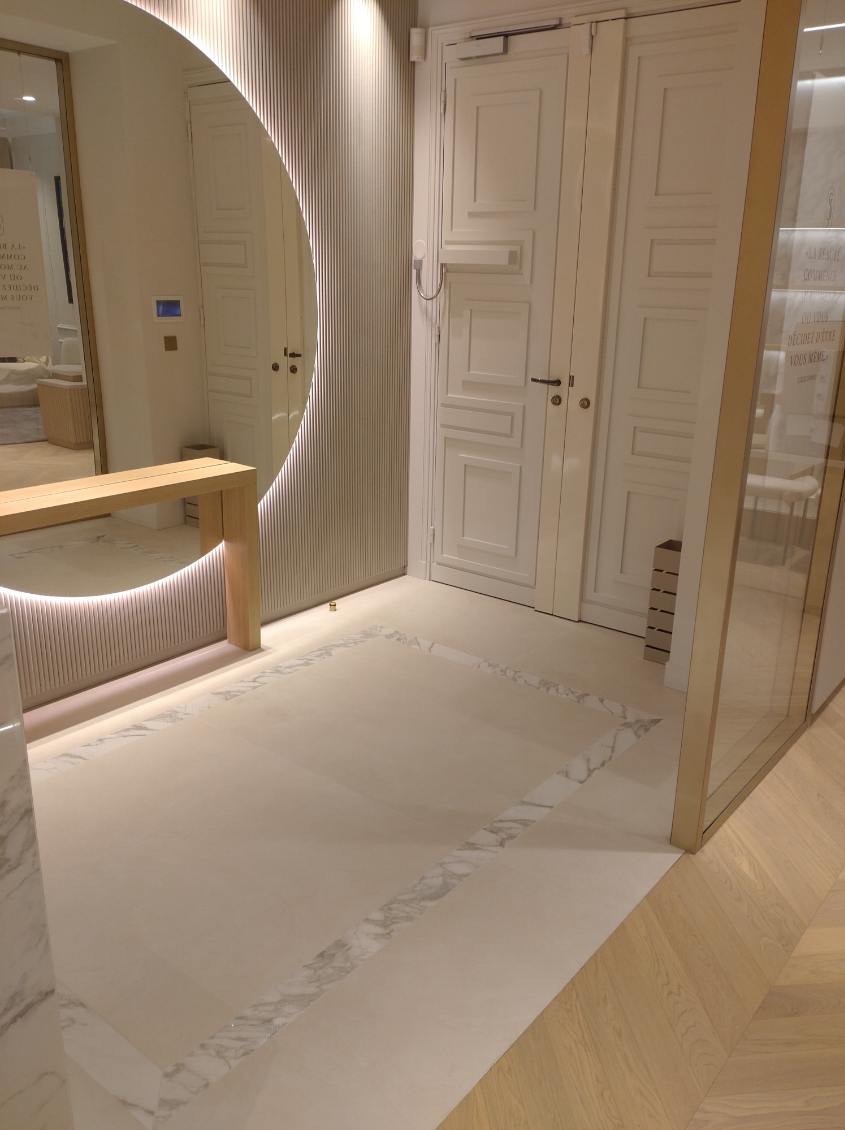 Nowoczesna łazienka z beżowymi płytkami podłogowymi obramowanymi marmurowym wzorem, duże okrągłe lustro z podświetleniem LED i białe drzwi w tle.