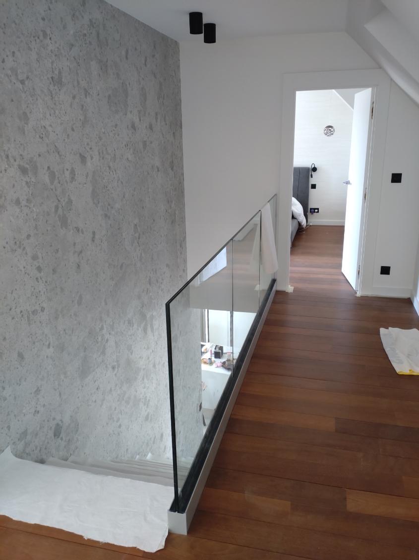 Korytarz z drewnianą podłogą i szklaną balustradą, ściana pokryta tapetą w odcieniach szarości, widok na otwarte drzwi do sypialni.