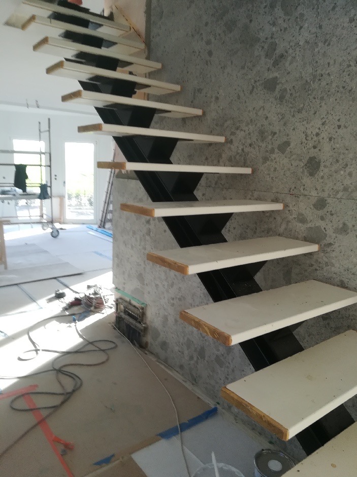 Nowoczesne schody z białymi stopniami i czarną, zygzakowatą konstrukcją nośną, w trakcie montażu przy ścianie wykończonej szarym kamieniem. W tle widoczne narzędzia i elementy wyposażenia wnętrza.