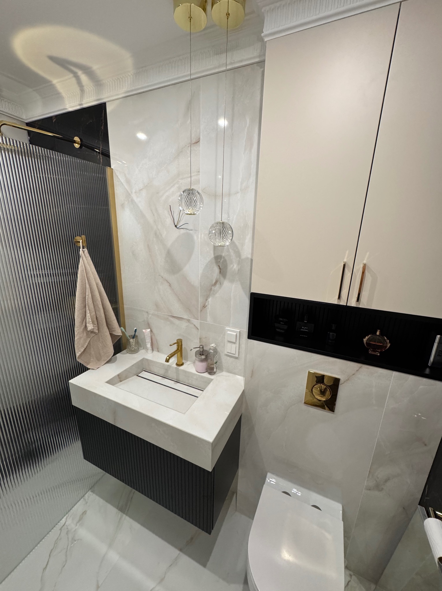 Elegancka łazienka z marmurowymi płytkami, wiszącą szafką z umywalką i złotymi akcentami, oświetlona wiszącymi lampami i z zabudową nad toaletą.