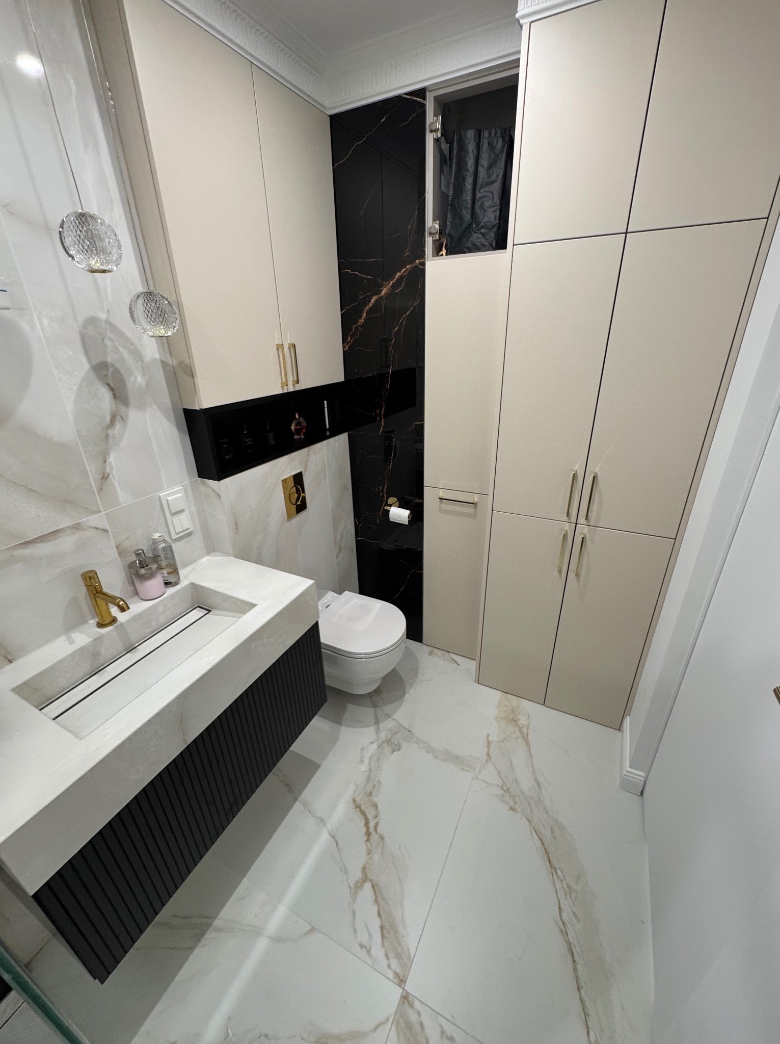 Elegancka łazienka z marmurowymi płytkami, beżowymi szafkami i złotymi akcentami. Umywalka z szafką w kolorze czarnym, wisząca toaleta i dekoracyjne lampy.