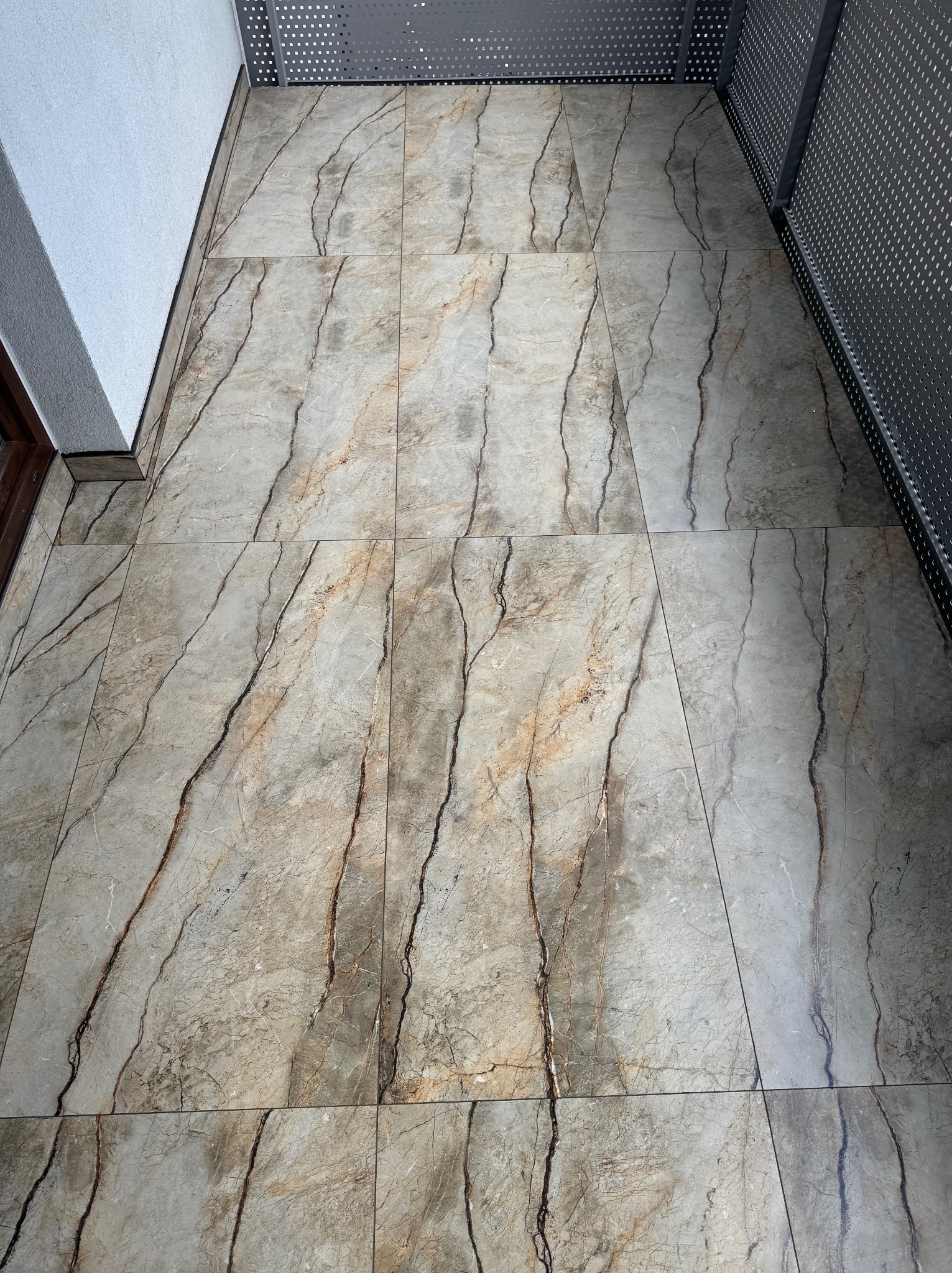 Ułożone płytki podłogowe imitujące marmur na balkonie, widok z góry, z fragmentem metalowej balustrady i ściany. Realizacja glazurnicza.