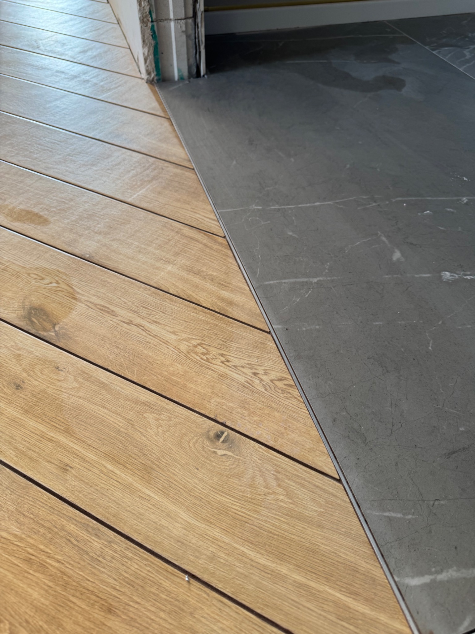 Połączenie parkietu ułożonego w jodełkę z szarymi płytkami w przejściu. Widoczny fragment progu. Jasne drewno kontrastuje z ciemnym kamieniem.