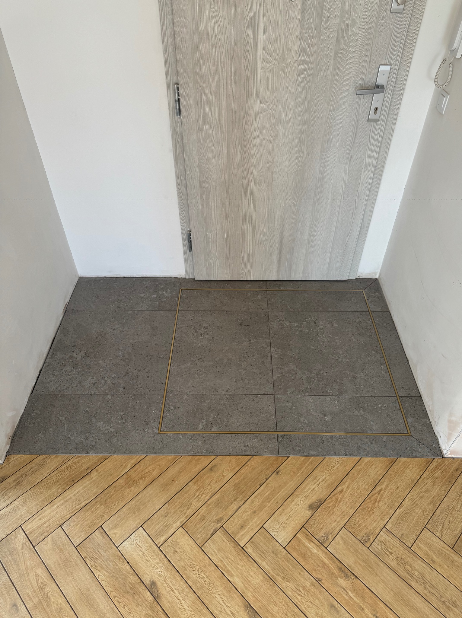 Szare płytki podłogowe z metalowymi listwami przy drzwiach, przechodzące w parkiet ułożony w jodełkę. Świeżo wykończone ściany w przedpokoju.