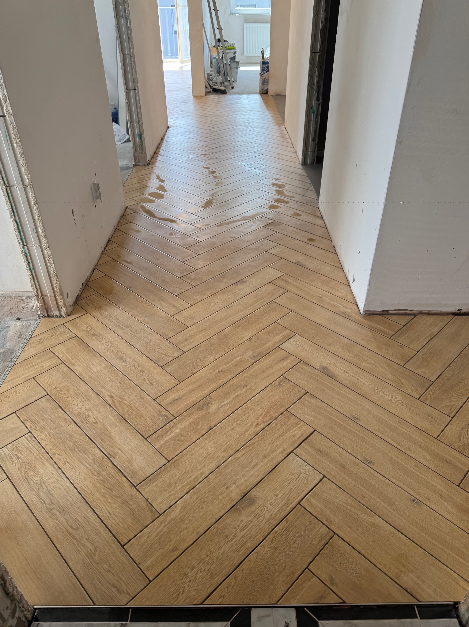 Ułożona jodełka z paneli podłogowych w korytarzu, widoczne ślady wilgoci. W tle maszyna do szlifowania i niezakończone wykończenia ścian.