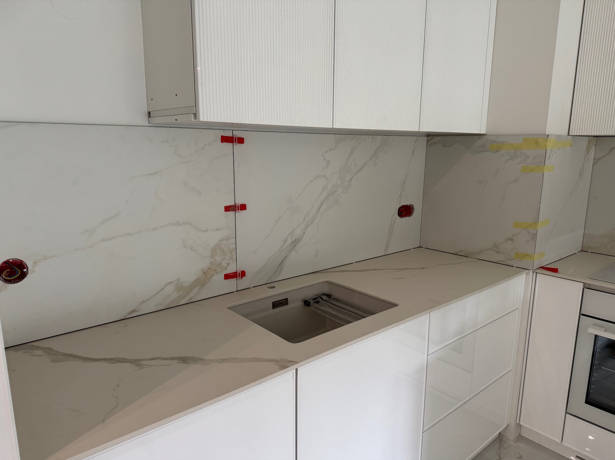Biała kuchnia w trakcie montażu: marmurowe płytki na ścianie z czerwonymi krzyżykami dystansowymi, blat z wbudowanym zlewem i białe szafki wiszące.