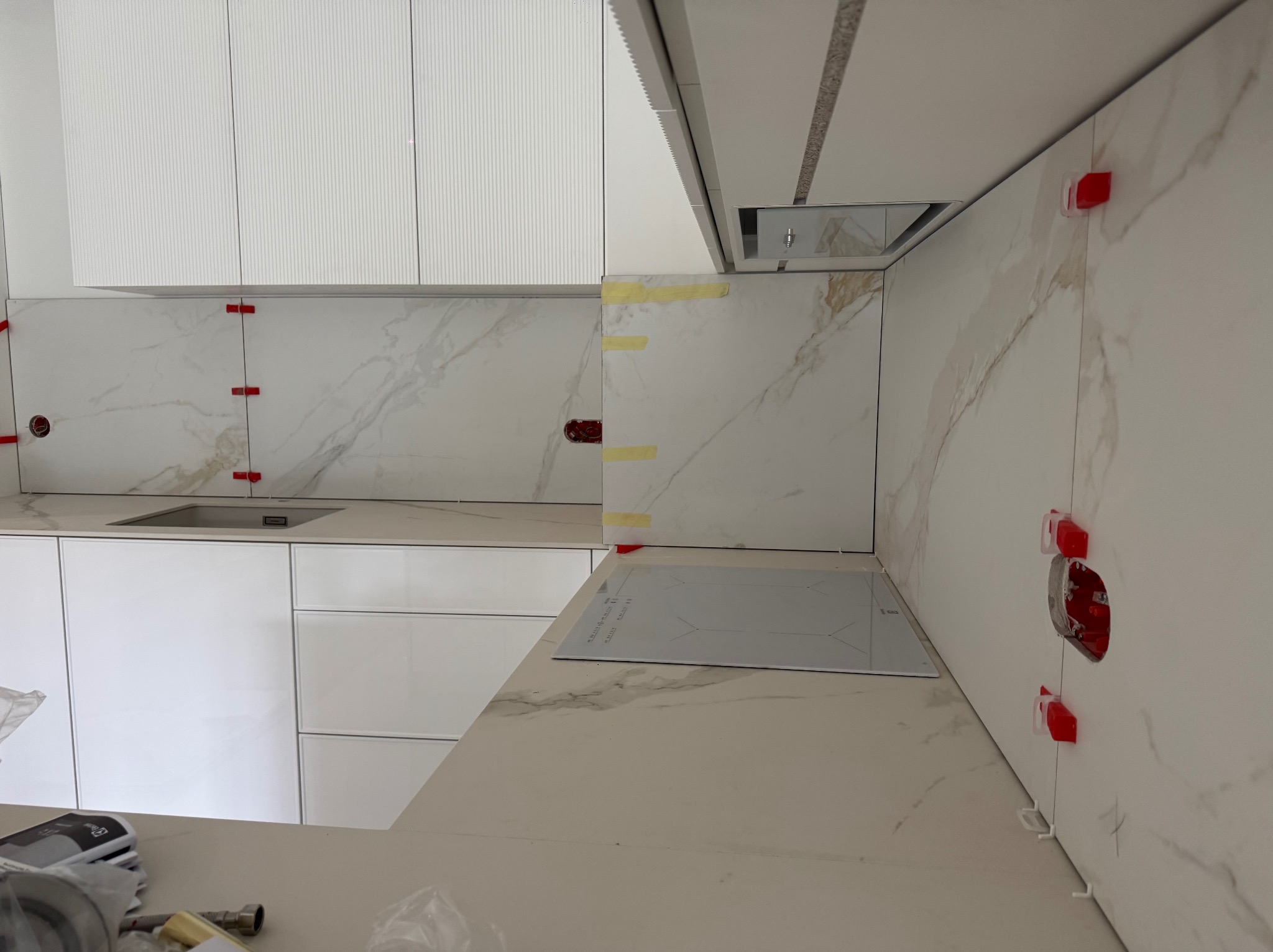 Białe płytki imitujące marmur na ścianie w kuchni, z czerwonymi klipsami poziomującymi. Widoczna płyta indukcyjna i białe szafki.