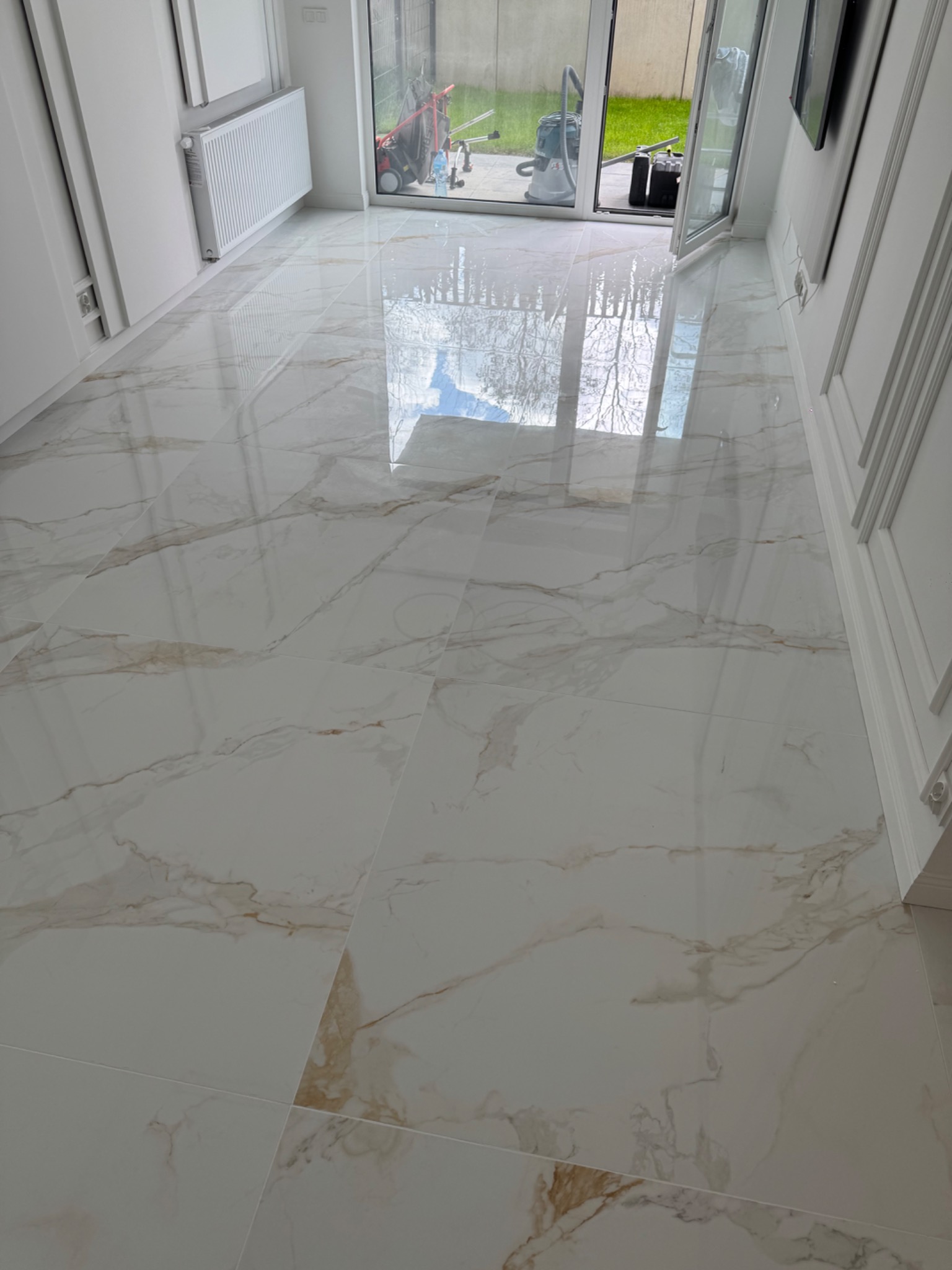 Błyszcząca, marmurowa podłoga w jasnym holu z widokiem na ogród przez przeszklone drzwi. Widoczny odkurzacz i narzędzia na zewnątrz.