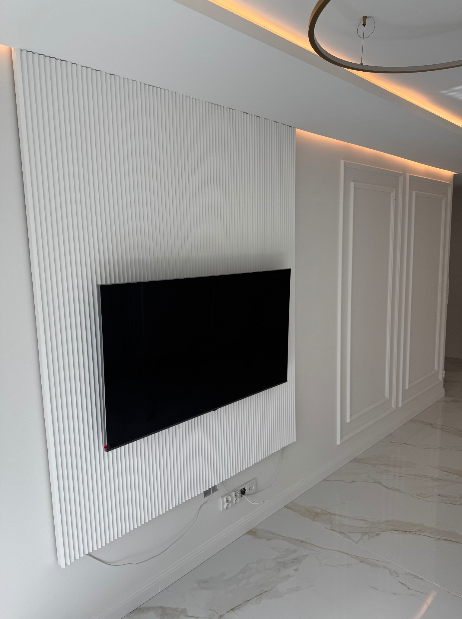 Nowoczesna ściana TV z białymi lamelami i czarnym telewizorem. Minimalistyczny design, ciepłe oświetlenie sufitowe, marmurowe płytki podłogowe.