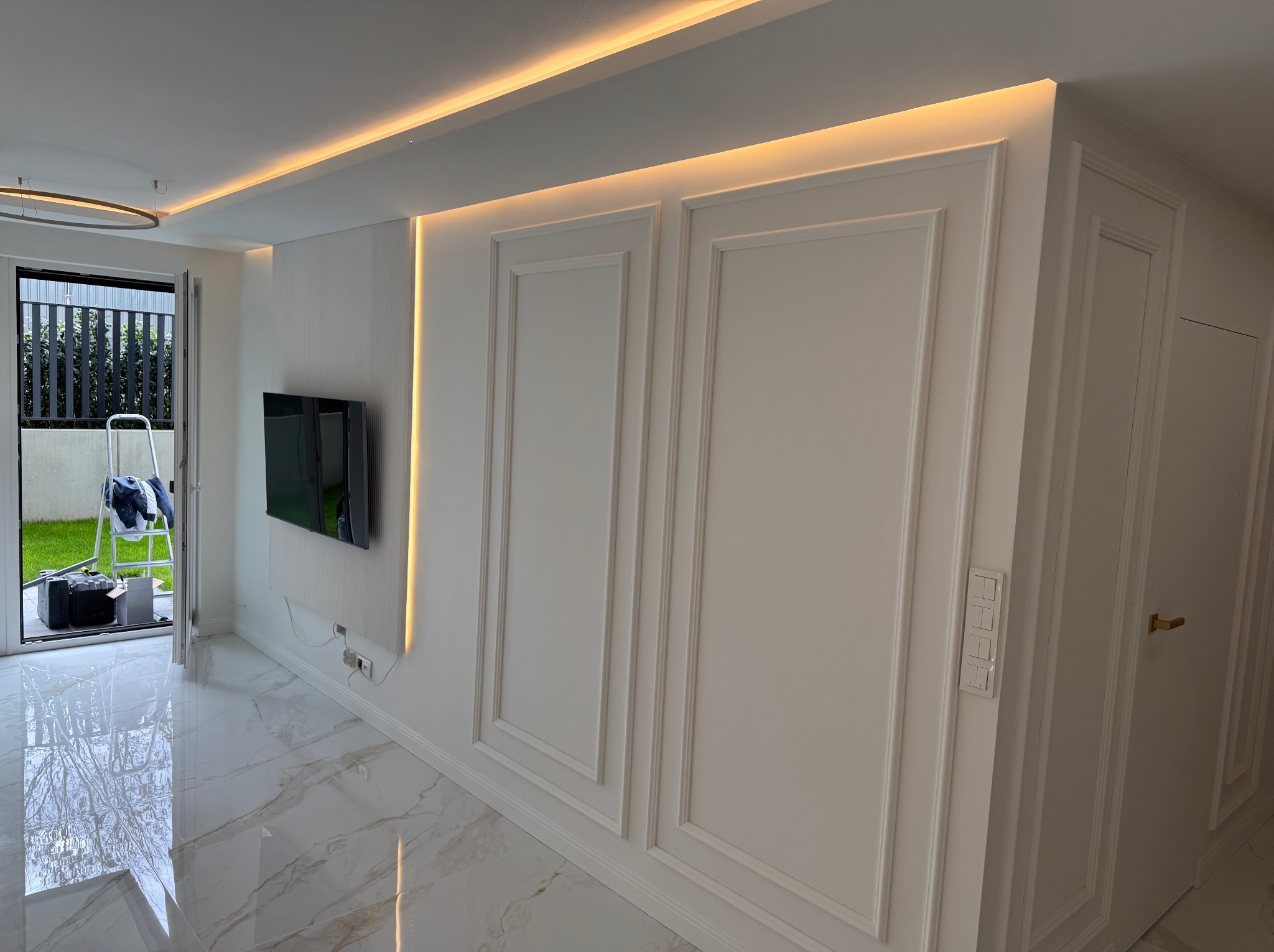 Eleganckie wykończenie wnętrza: białe ściany z dekoracyjnymi panelami, subtelne oświetlenie LED i marmurowa podłoga. W tle widoczne narzędzia malarskie.