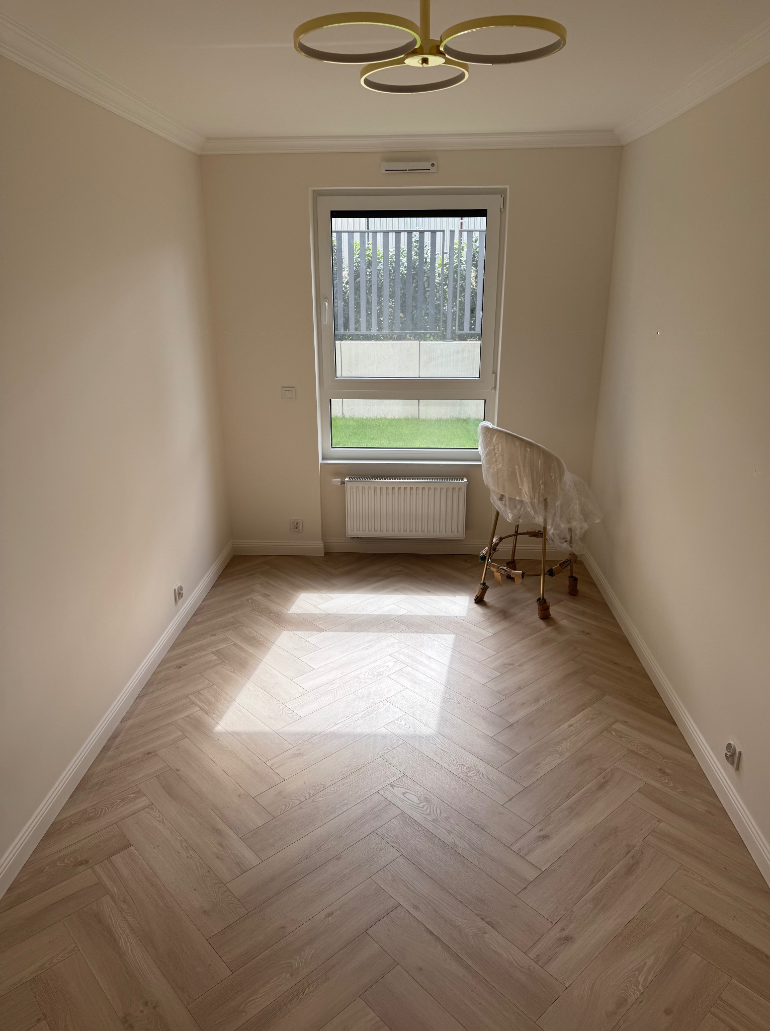 Jasne panele ułożone w jodełkę, kremowe ściany z listwami przysufitowymi, okno z widokiem na zieleń, złoty żyrandol. Krzesło zabezpieczone folią. Nowoczesne wykończenie wnętrza.