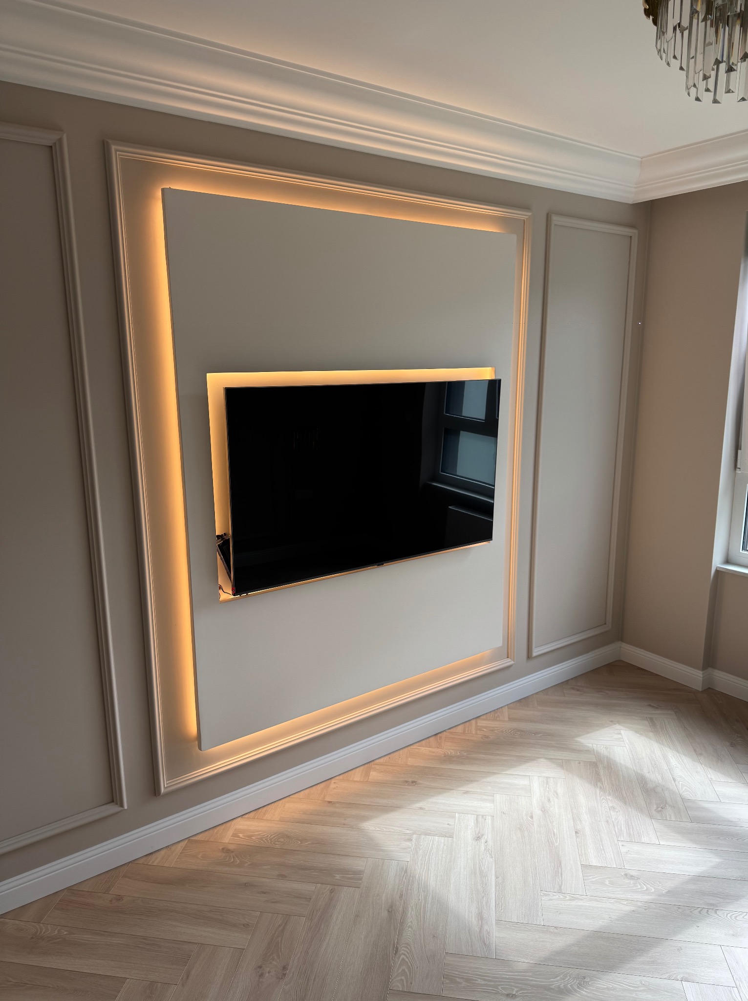 Nowoczesna ściana z wnęką na TV, podświetlona LED, z dekoracyjnymi listwami i parkietem ułożonym w jodełkę. Minimalistyczny design wnętrza w jasnych, stonowanych barwach.