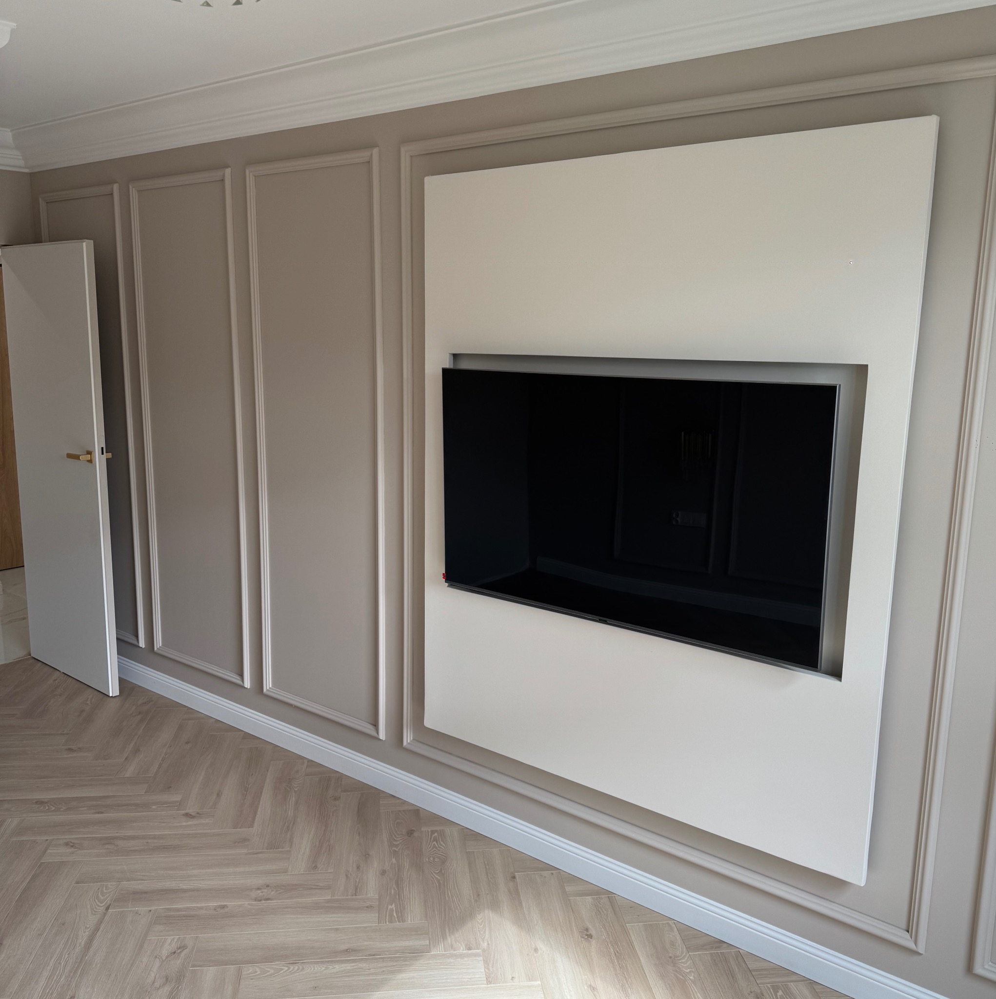 Eleganckie wykończenie salonu: ściana z dekoracyjnymi panelami w odcieniach beżu, wbudowany telewizor, parkiet w jodełkę, otwarte drzwi. Klasyczny design.