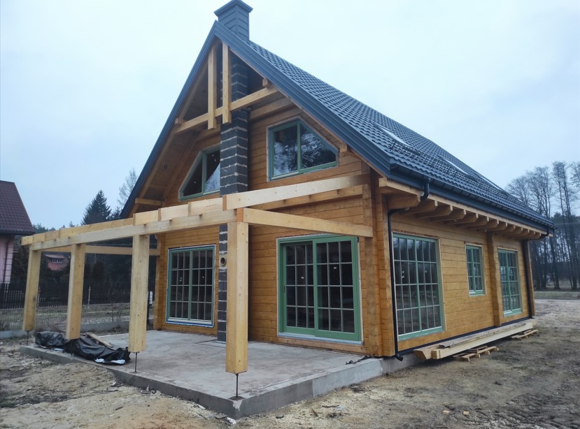 Surowy stan domu z drewnianych bali z widoczną konstrukcją zadaszenia tarasu, zielone ramy okienne, czarny dach i komin z cegły klinkierowej.