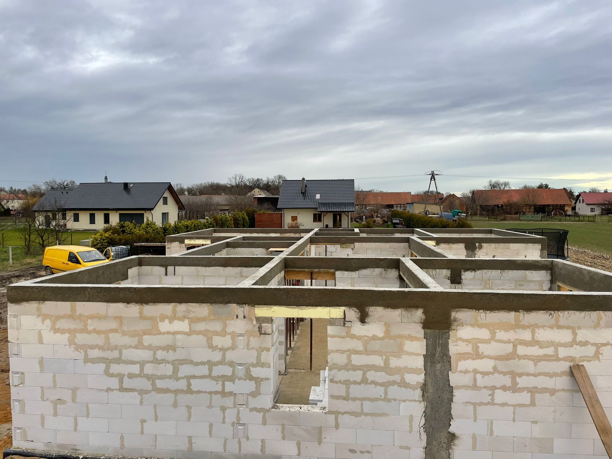 Wznoszenie ścian parteru z betonu komórkowego na fundamencie, widok z góry na budowę domu jednorodzinnego z żółtym samochodem dostawczym w tle.