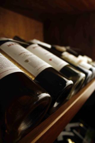 Rząd butelek wina ułożonych na drewnianej półce w piwniczce, etykiety częściowo widoczne, ciemne szkło butelek.