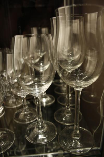 Kieliszki do wina o różnych kształtach i rozmiarach, ustawione w rzędach na szklanej półce, widoczne refleksy światła na szkle.