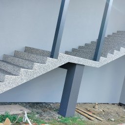 KWwork Kacper Wróbel - Zewnętrzne schody pokryte szarym dywanem kamiennym, wsparte na ciemnoszarych słupach, przylegające do jasnoszarej ściany budynku.