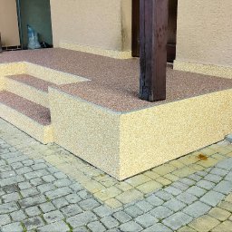 KWwork Kacper Wróbel - Zewnętrzne schody wejściowe do budynku pokryte beżowo-brązowym dywanem kamiennym, z widocznymi dwoma stopniami i drewnianymi kolumnami podtrzymującymi zadaszenie, na tle kostki brukowej.