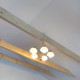 KWwork Kacper Wróbel - Poddasze z widocznymi drewnianymi belkami konstrukcyjnymi i wiszącą lampą z pięcioma mlecznymi kloszami, częściowo widoczne okno w czerwonej ramie.