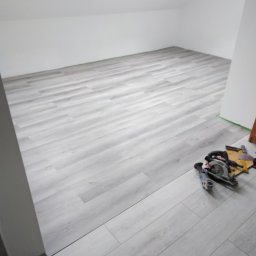 KWwork Kacper Wróbel - Jasnoszara podłoga panelowa ułożona w pokoju na poddaszu, widoczne narzędzia do cięcia paneli oraz komoda w rogu pomieszczenia.