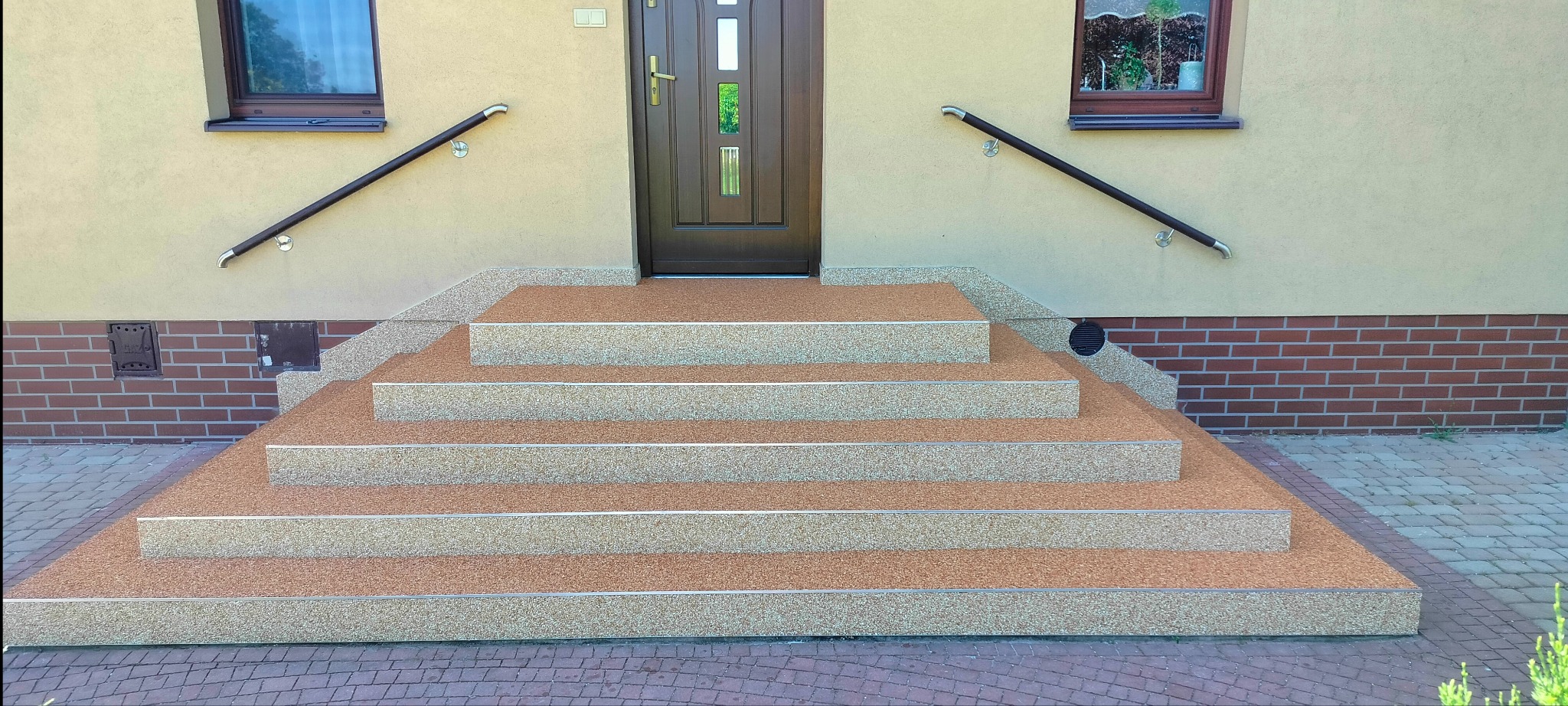 Schody wejściowe do domu wykończone żywicą epoksydową z kruszywem kwarcowym, tworzące wzór dywanu kamiennego, z metalowymi listwami na krawędziach stopni.