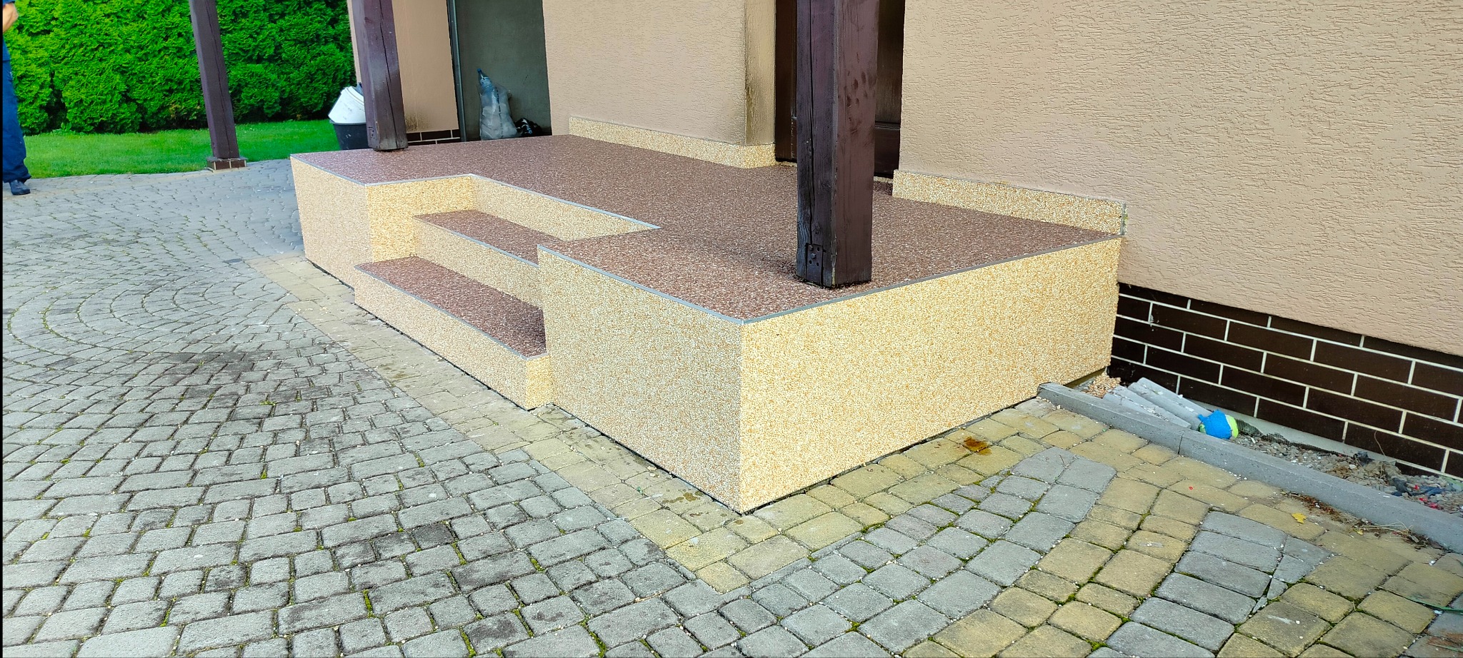 Zewnętrzne schody wejściowe do budynku pokryte beżowo-brązowym dywanem kamiennym, z widocznymi dwoma stopniami i drewnianymi kolumnami podtrzymującymi zadaszenie, na tle kostki brukowej.