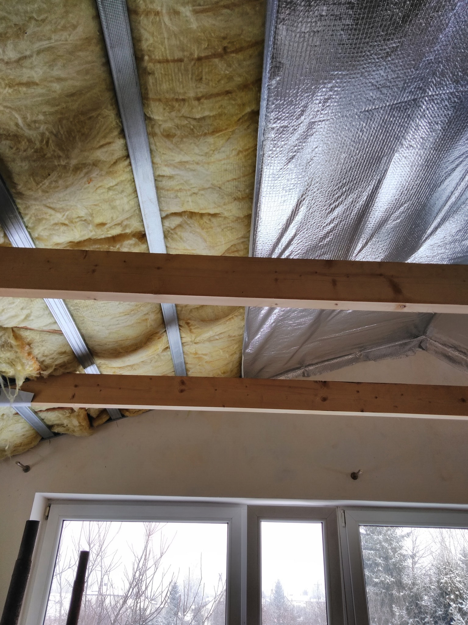 Poddasze w trakcie ocieplania: widoczna wełna mineralna, folia aluminiowa oraz metalowe profile konstrukcyjne, drewniane belki i okno z widokiem na zimowy krajobraz.